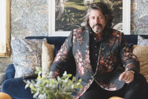 A man wearing a black floral suit sits on a sofa in front of a picture and patterned wallpaper