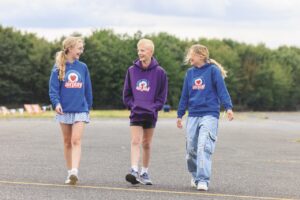 Three children wearing purple and blue hoodies walk in a row across a playground. They smile and laugh with each other