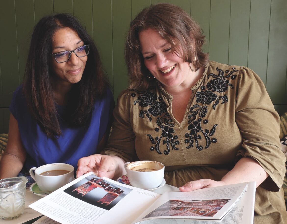 Two women sit at a table with coffee, reading a book in front of them and smiling