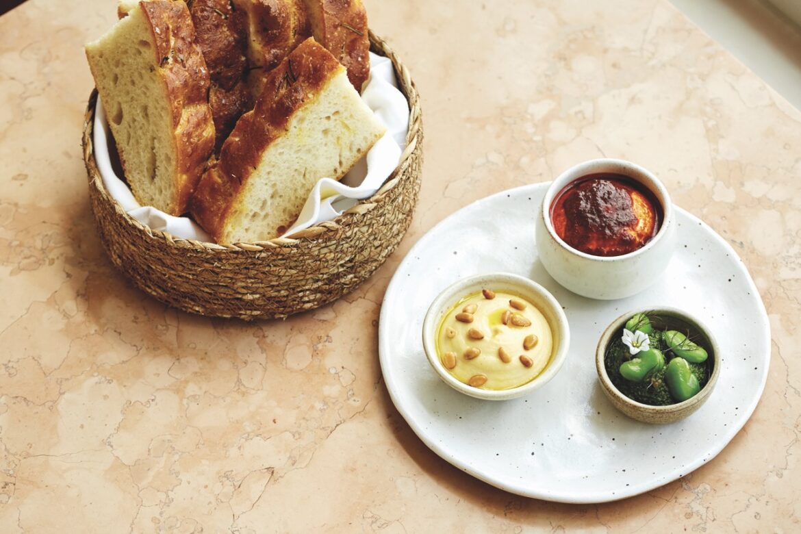 Top view of a basket of sliced bread next to a plate with three different dips in small bowls