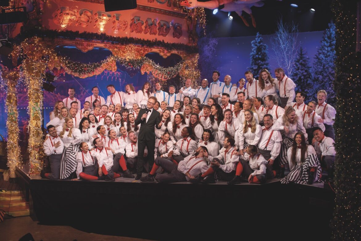A group of singers wearing white shirts standing onstage with a Christmas backdrop. A man in a suit with a microphone stands in the centre