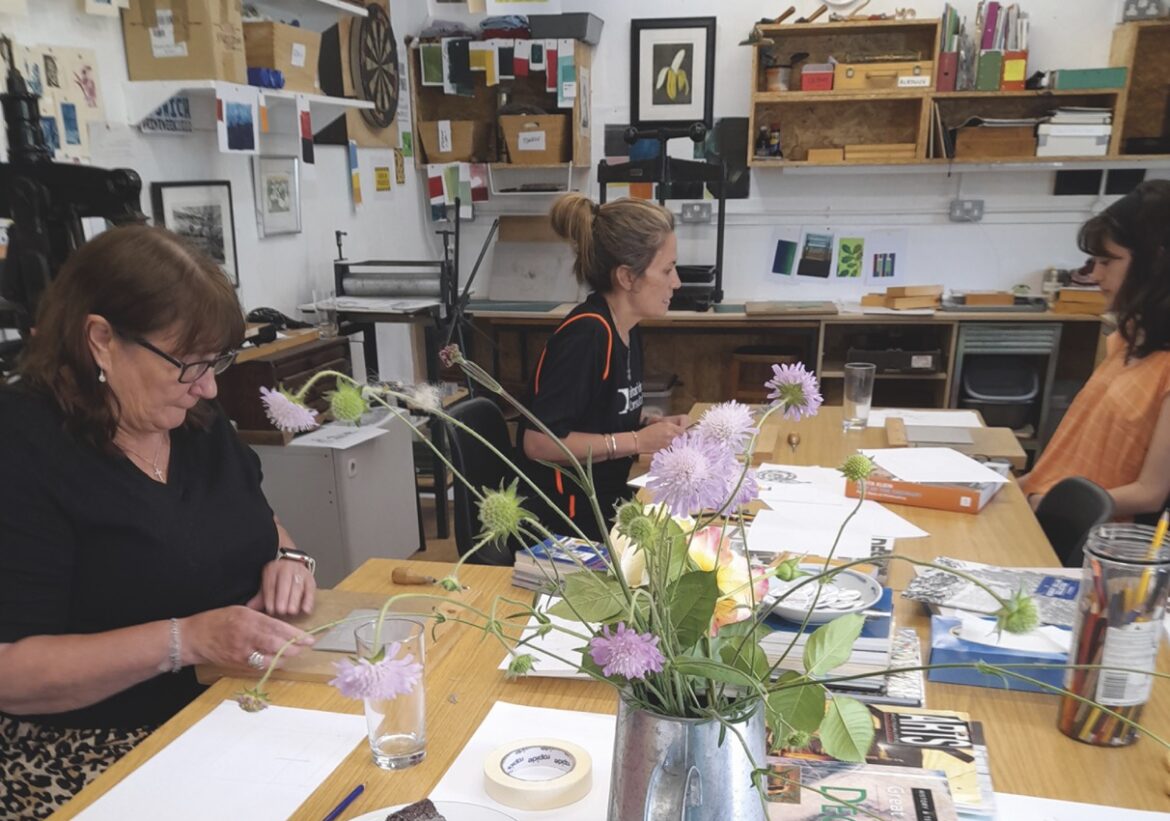 Three women sit around a table in an art workshop, working on prints