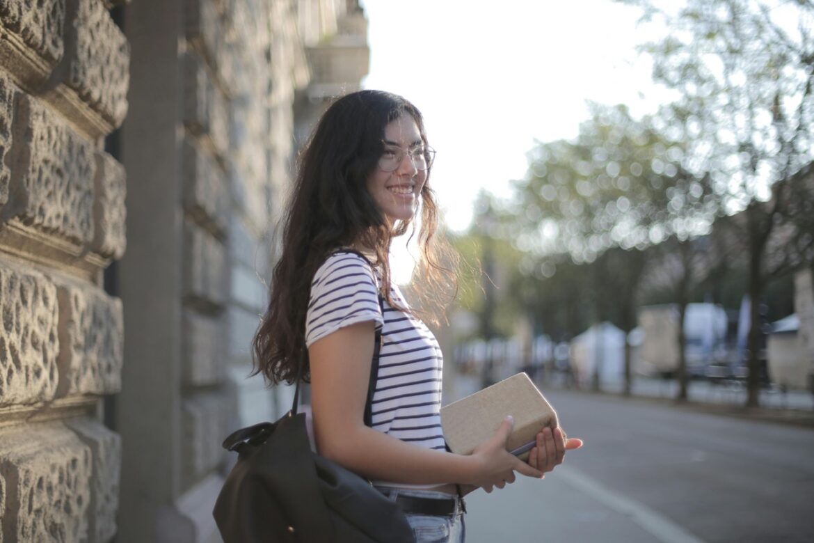 A young woman stands on a street holding a book in her hands while smiling