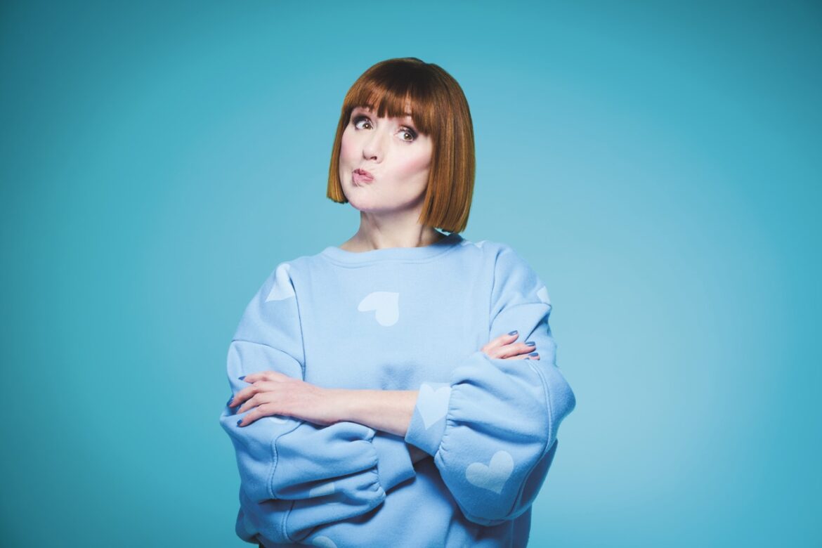 A woman with a short bob stands with her arms crossed, wearing a light blue jumper against a blue background