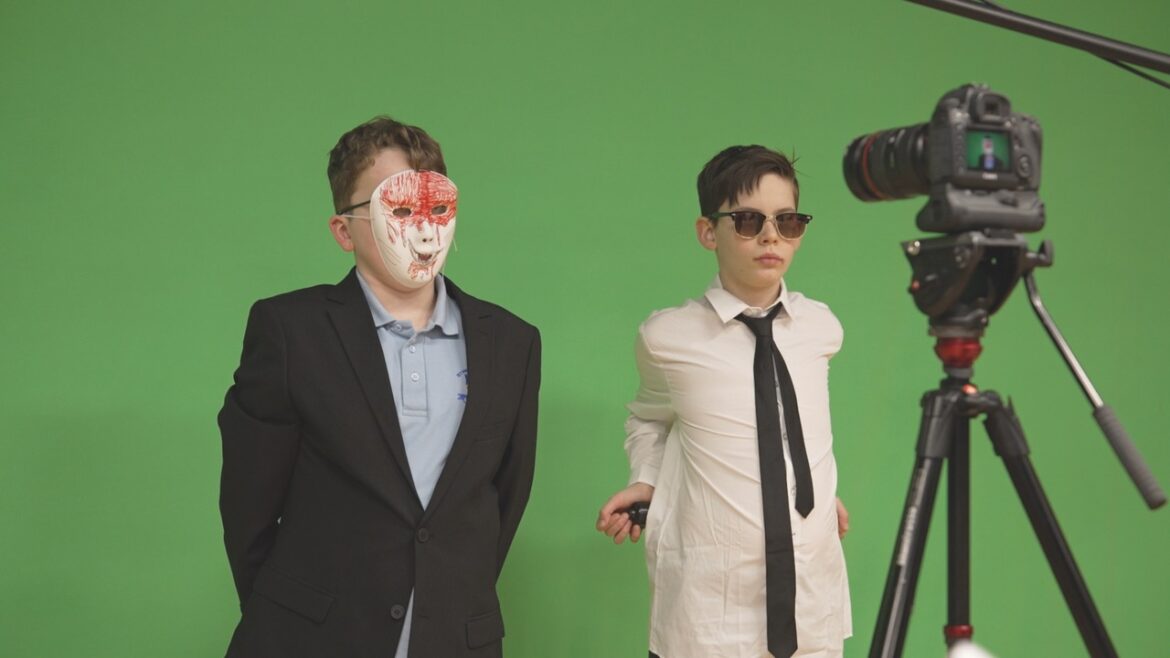 Two young children standing in front of green screen and looking at a professional camera