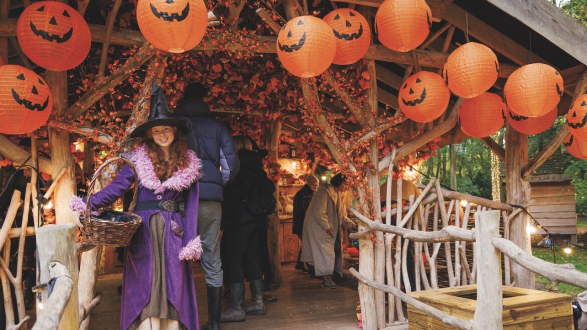 A young girl dressed in a purple witch outfit standing outside an autumnal grotto. She holds a basket and stands under pumpkin lanterns