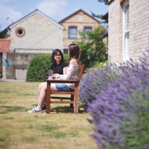 Two young girls sit on a wooden bench next to a lavender bush