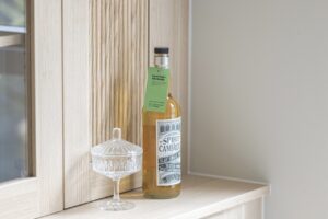 A bottle of orange liquid with a green label around the neck. It is placed next to a glass tableware on a sideboard