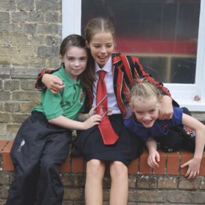 Three young girls in uniform sit with their arms around each other while sitting on a wall