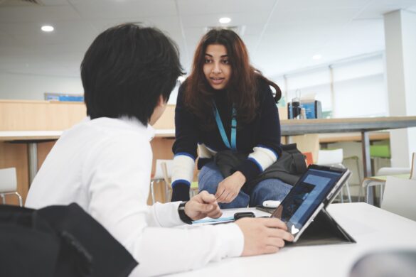 Two students talking in a common room. The boy sits at a table with a laptop open in front of him