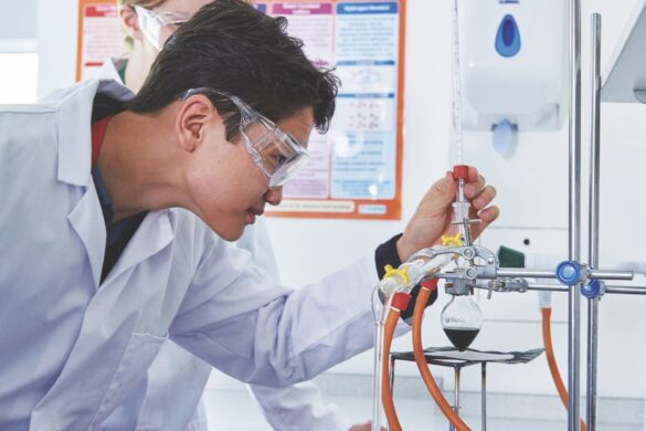 A sixth form student adjusting a flask in a school laboratory while wearing a white coat and safety goggles