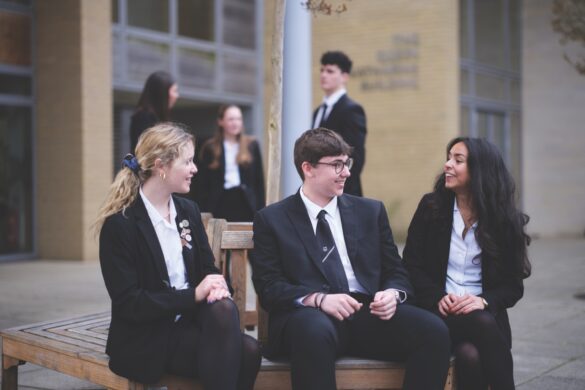 Three sixth form students sitting on a bench and talking, while more students stand behind