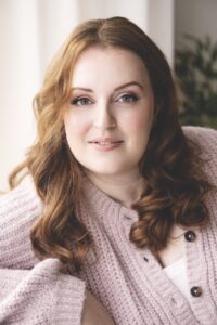 A young woman with curled hair and a pink cardigan sitting on a sofa in front of a white curtain