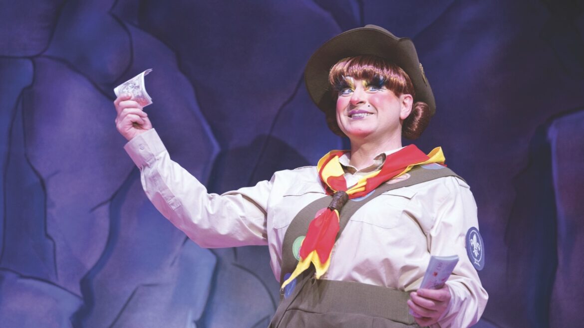 A pantomime dame standing in scout uniform and holding a piece of paper on a stage