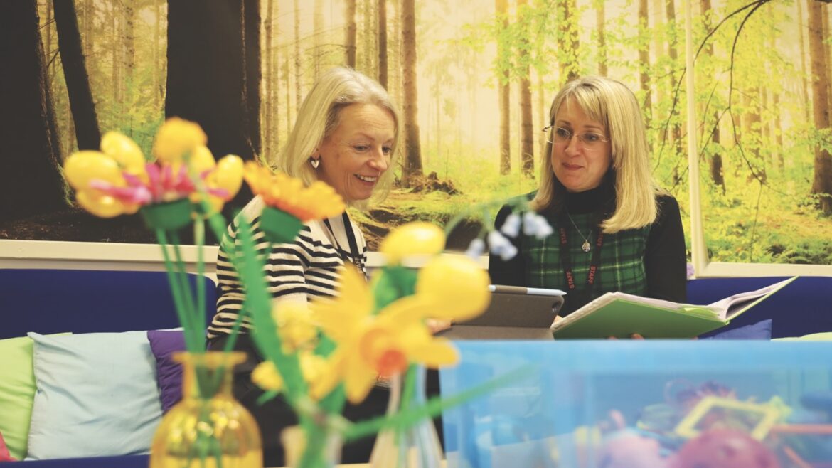 Two women looking at a tablet and sitting on a couch in a yellow room with toy flowers in the foreground