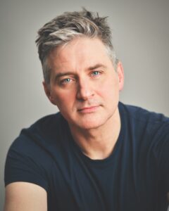 A man wearing a black t-shirt in front of a grey background