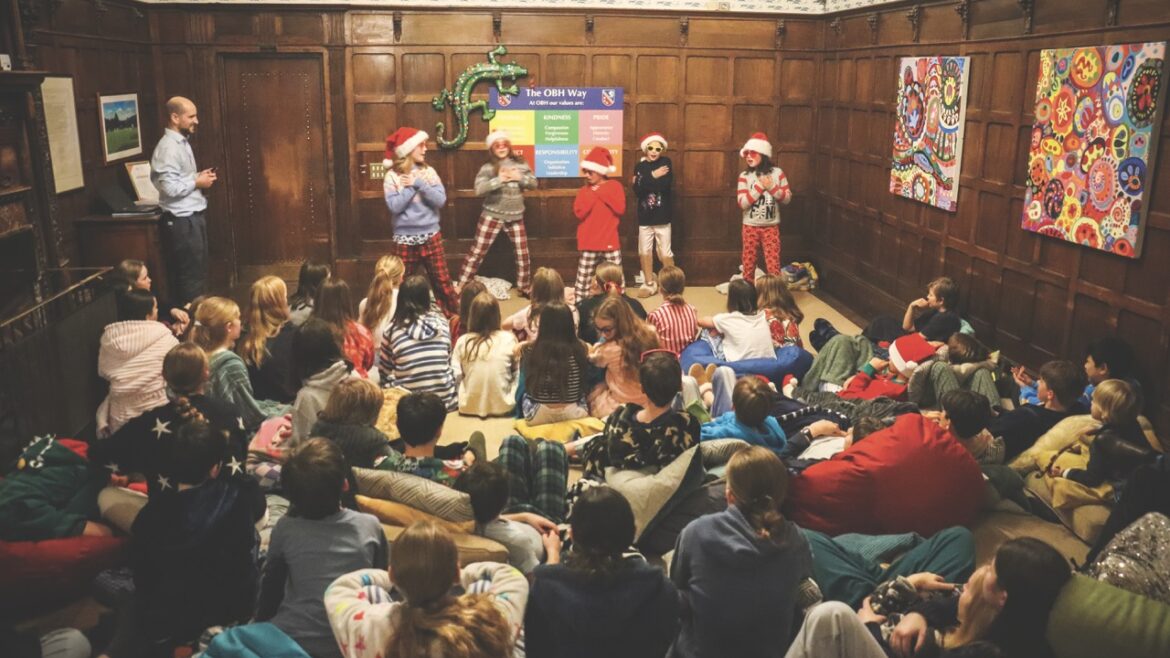 Children gathered in an ornate room. They sit on the floor and watch five children in christmas pyjamas as they perform at the front