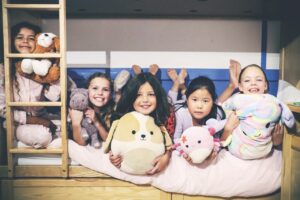 Five children sit on a bunkbed holding stuffed animals and pillows