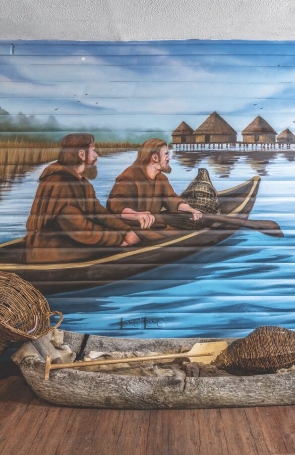 A painting of people in a boat with a real log boat placed in front in a museum