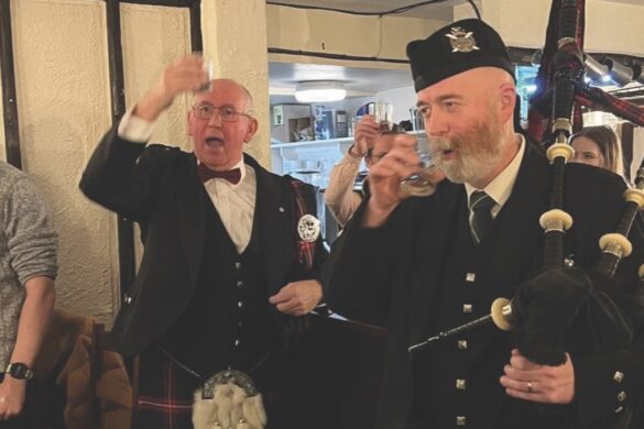 Two men stand in kilts in a pub while they drink beer