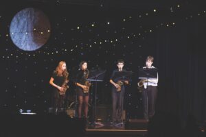 A group of four saxophone players standing on a stage with a space background
