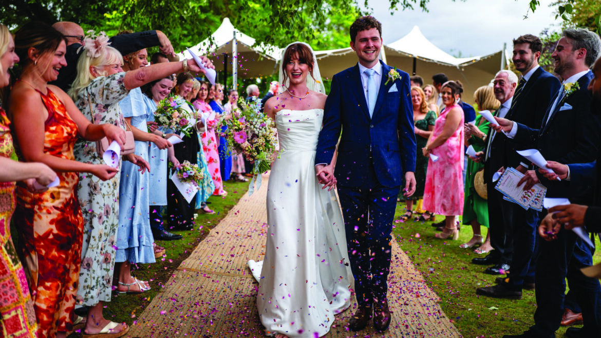 A bride and groom walking down an outside aisle as people either side throw confetti at them