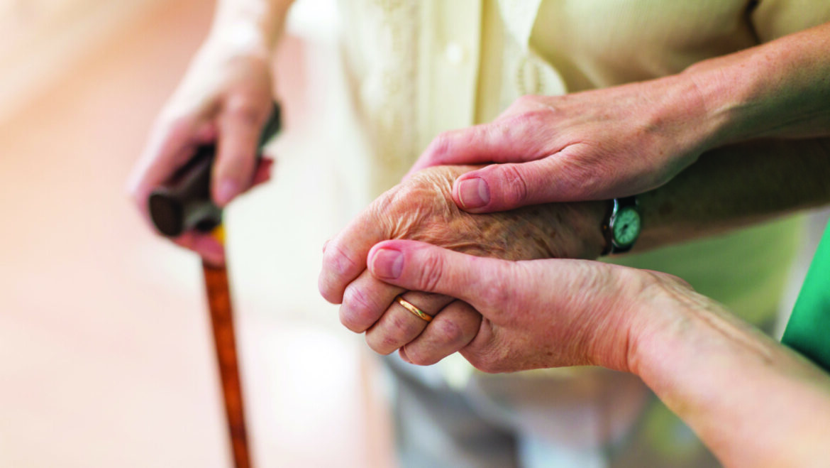 A pair of hands holding an older hand wearing a wedding ring and watch. Another hand in the blurred background holds a walking stick