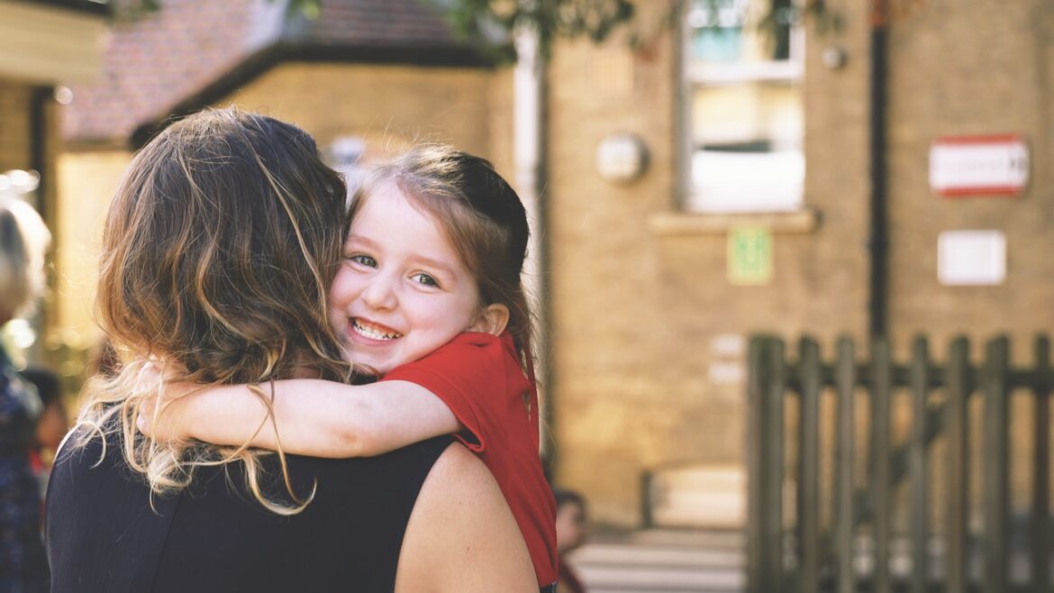 A young girl in a red top smiling at the camera as she hugs a woman with her back to the camera