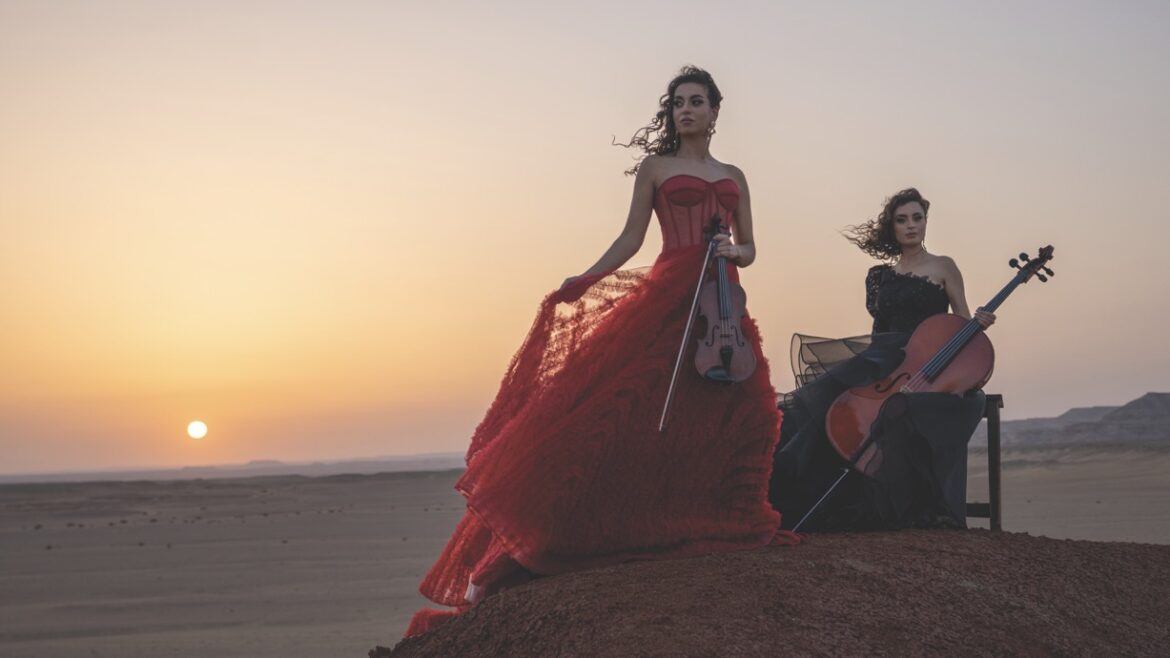 Two women in dresses holding instruments as they stand at the top of a hill at sunset