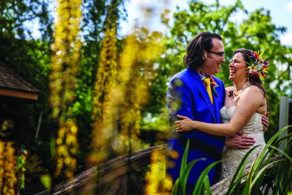 A bride and groom dressed in bright colours, embracing behind foliage