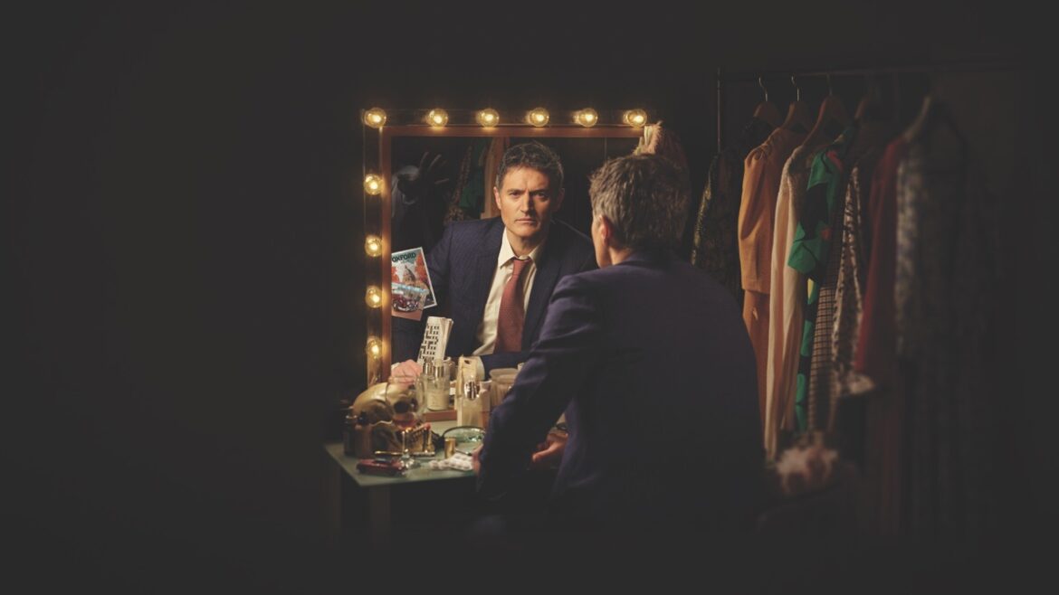 A man in a suit sitting in darkness, staring into a mirror with lightbulbs around the edge