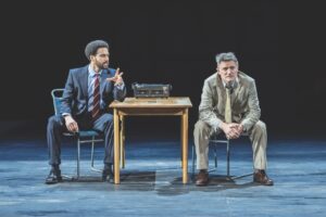 Two men sitting in suits sitting either side of a table on a stage