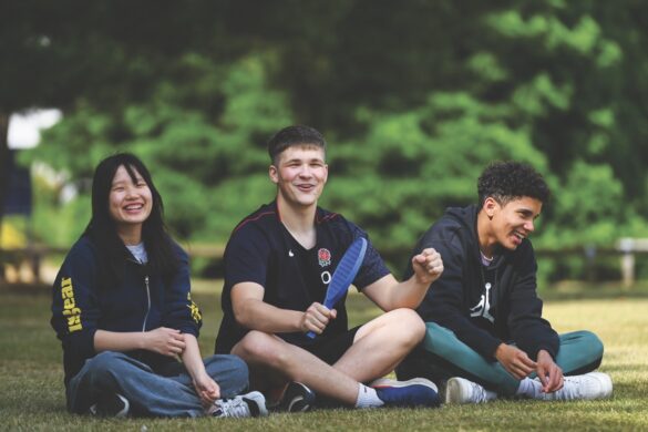 Students sitting on the grass in a row outside. One holds a blue bat