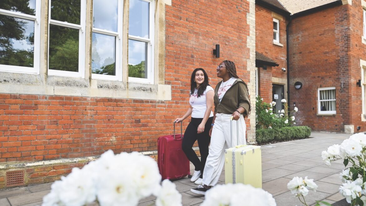 Two students walk with suitcases past flowers and a brick building