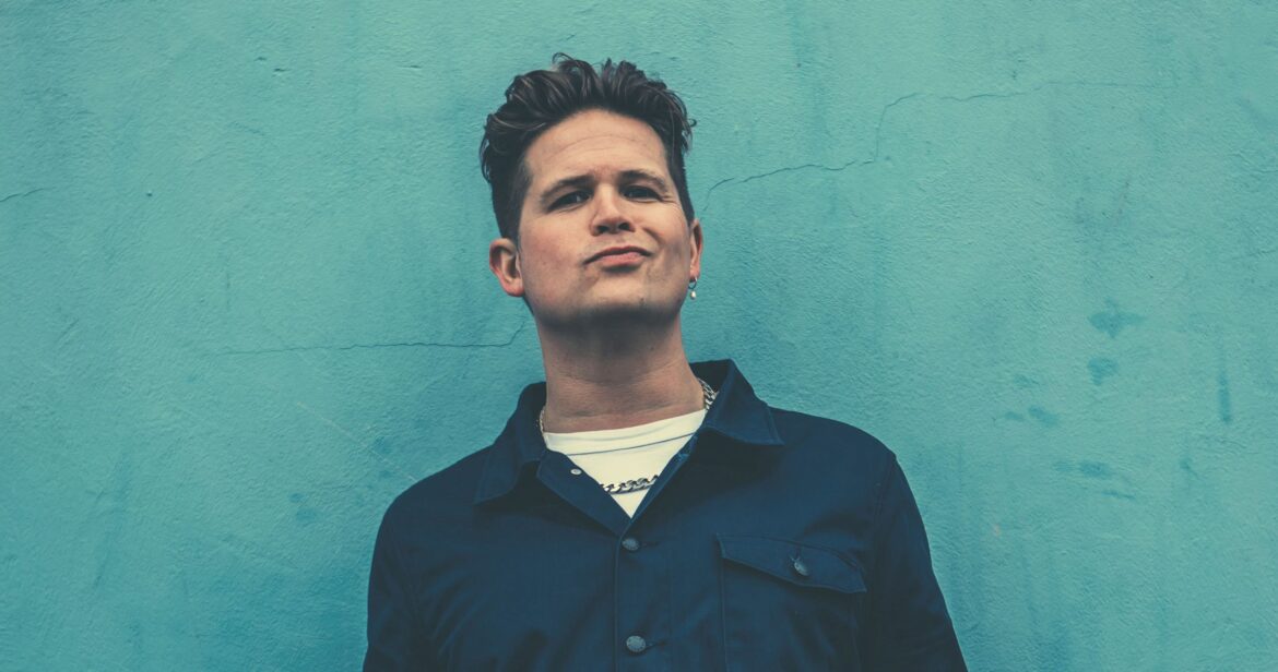 A man with a smirk leaning against a teal wall